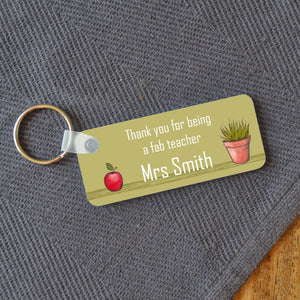 Personalised green key ring with a design which includes an illustration of an apple and a pot plant, and a message which reads "thank you for being a fab teacher Mrs Smith" 