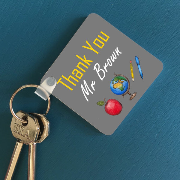 A square key ring on a blue table with a set of keys. The key ring has a personalised design which includes an apple, a globe a pen and a pencil. It is also customised with a message reading "Thank you Mr Brown"