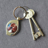 A personalised teardrop shaped metal keyring. The keyring is on a table. It has been personalised with a family photo. 
