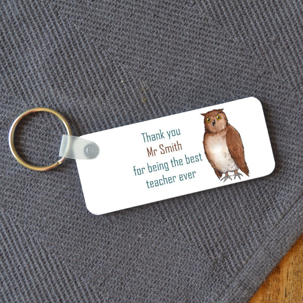 A rectangular key ring on a wooden table. The key ring has a personalised design printed on it which includes an illustration of an owl and a message reading "thank you Mr Smith for being the best teacher ever"