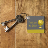 A personalised keyring on a table with a set of keys. The keyring is square and the design is grey and yellow. The design includes a yellow globe and the message "thank you Mr Smith"