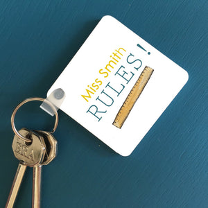 A square key ring on a blue table with a personalised design. The design includes a message which reads "Miss Smith Rules!" and an illustration of a ruler. The text is yellow and teal. 