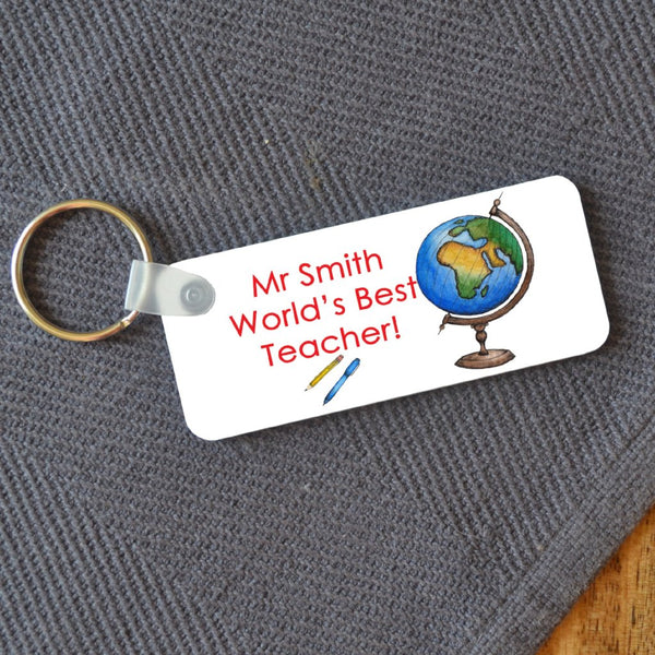 A rectangular key ring on a table which has a personalised design on it. The design includes an illustration of a globe and the message "Mr Smith, worlds best teacher"
