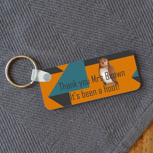 A rectangular keyring on a table with a personalised design. The design includes an illustration of an owl and a message reading "thank you mrs brown, its been a hoot!" the design is on an orange, teal and grey background.